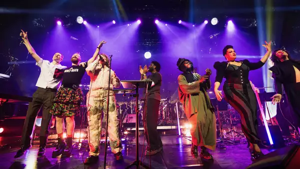 Multiple people on a stage singing, clapping and dancing with purple lighting