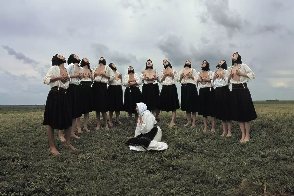 A group of women in traditional Eastern European attire stand barefoot in a grassy field under a dramatic, cloudy sky. Most are wearing black skirts, white blouses, and black headscarves, with some partially exposing their chests while lifting their heads upward in a ritualistic pose. In the foreground, a single woman dressed in white kneels on the ground with a headscarf and embroidered belt, gazing to the side.