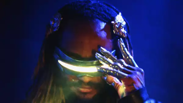 Thundercat wearing futuristic glowing visor glasses and metallic claw-like gloves against a dark blue background.