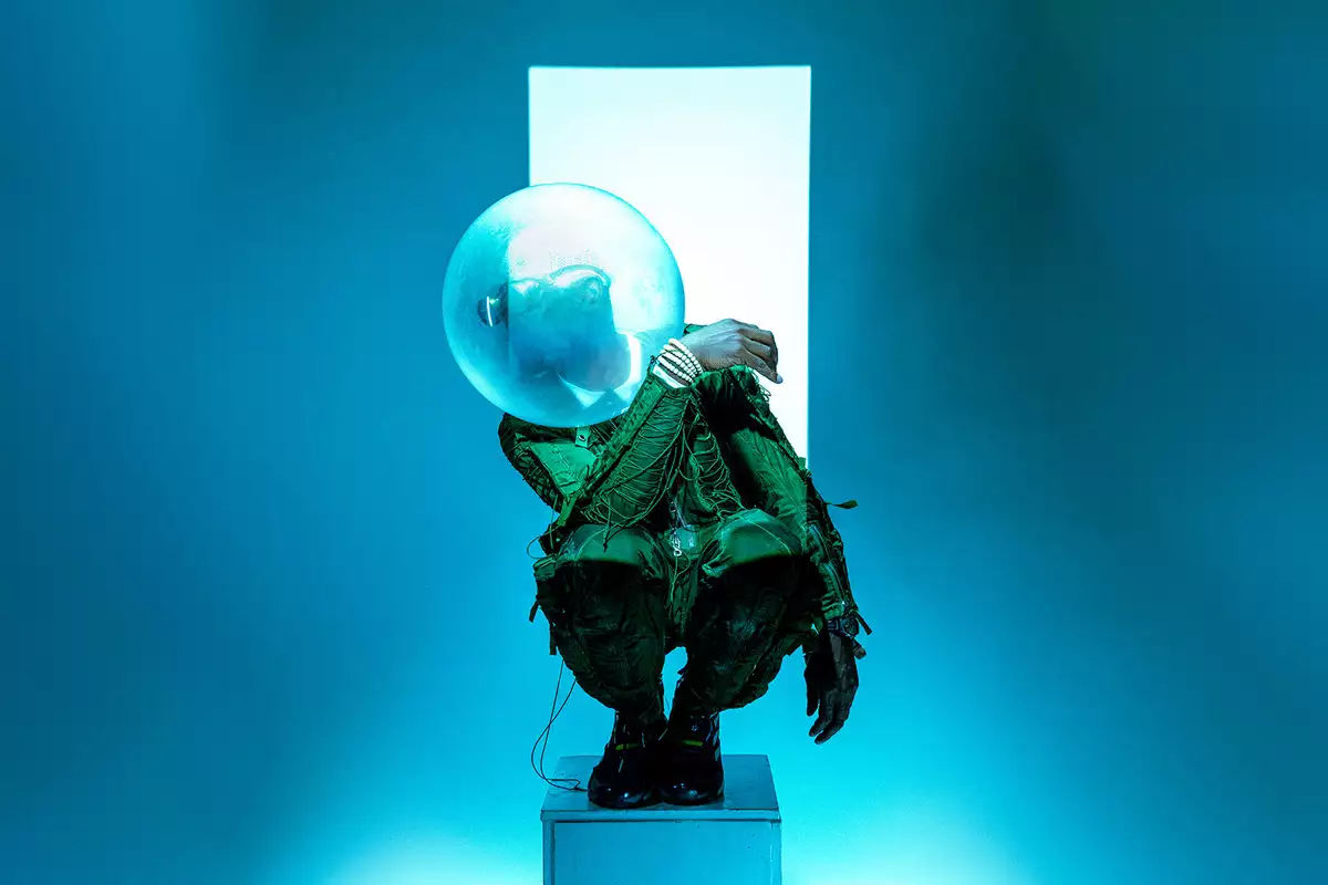Benji Reid crouched on a small pedastal in front of a white strip against a blue background. He is wearing a green pattered suit and a bubble sculpture on his head.