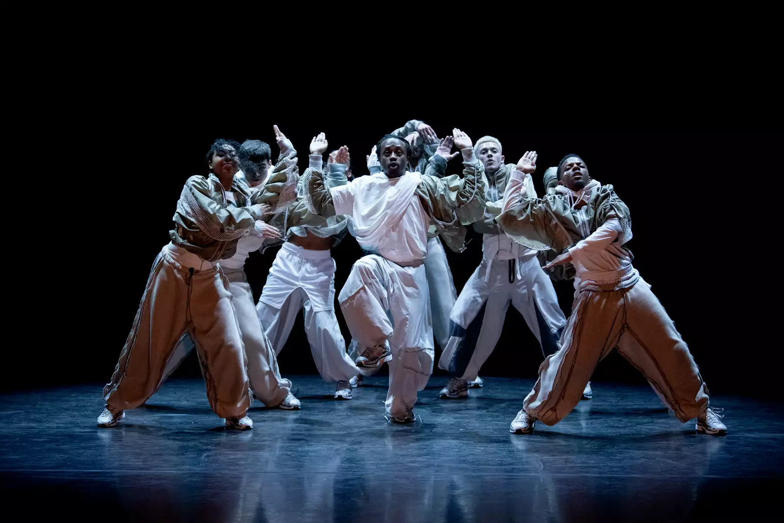 A group of dancers performing on a dark stage during CYCLES, wearing white clothing.