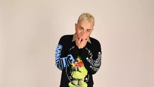 Joshua James stood against a beige background, with their hands raised over their nose