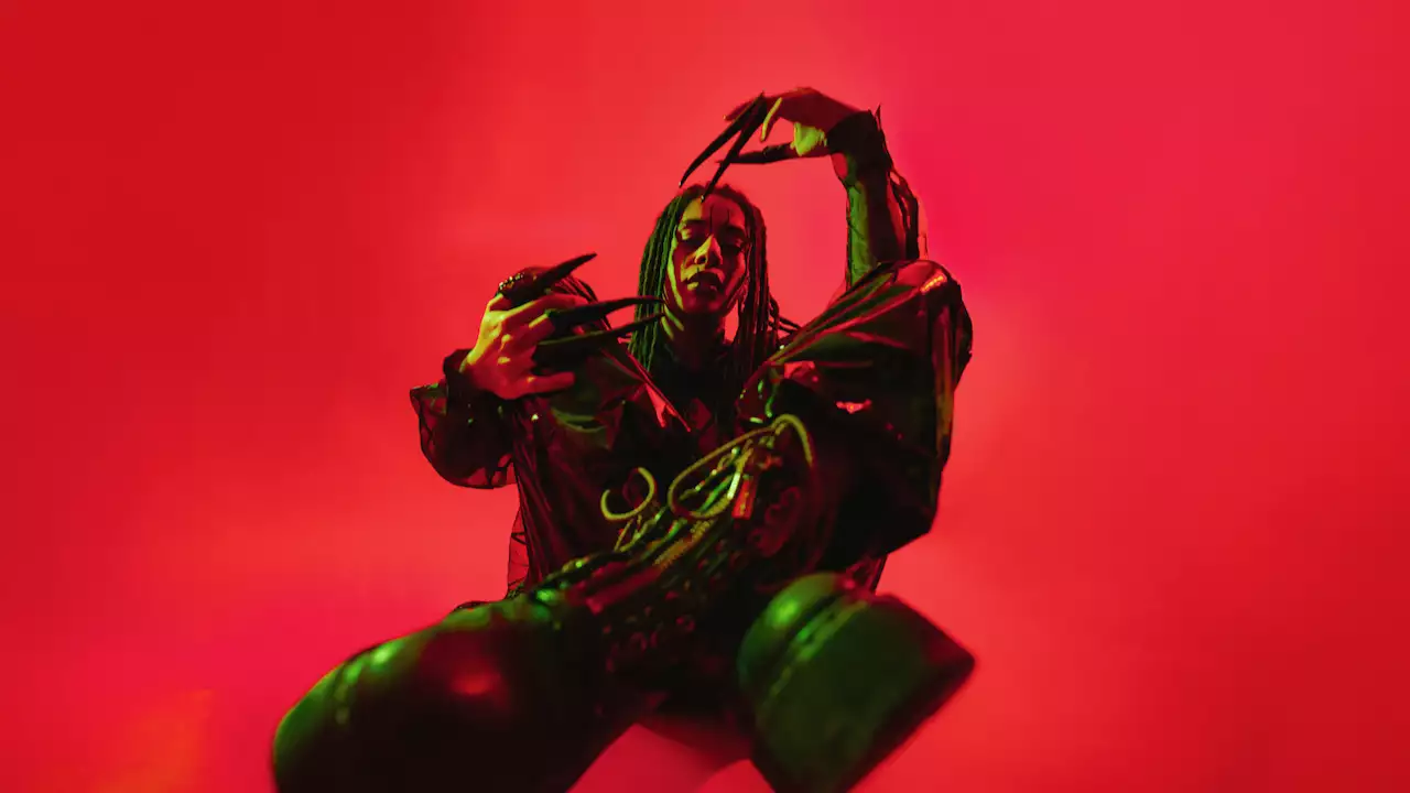 GROVE wearing all black and long black nails, sat crossed legs on a bright red background