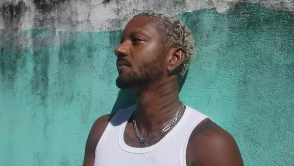 Atike stands in profile against a turquoise and white wall, wearing a white tank top and layered silver necklaces.