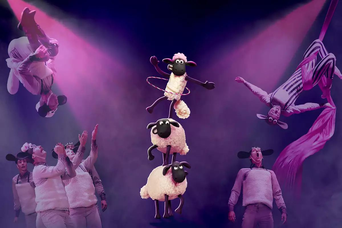 Performers in sheep costumes execute acrobatic stunts and aerial silk acts under dramatic pink lighting. In the center, three animated sheep from "Shaun the Sheep" balance in a tower, with the top one hula-hooping.