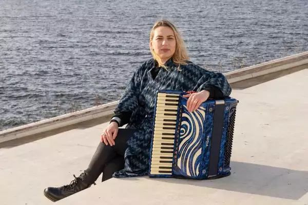 Photo of Tuulikki Bartosik sat by the water with her accordion