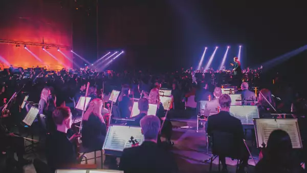 The BBC Philharmonic orchestra performing in the Warehouse. The lighting is pink and purple.  The audience are stood in the middle of the orchestra.