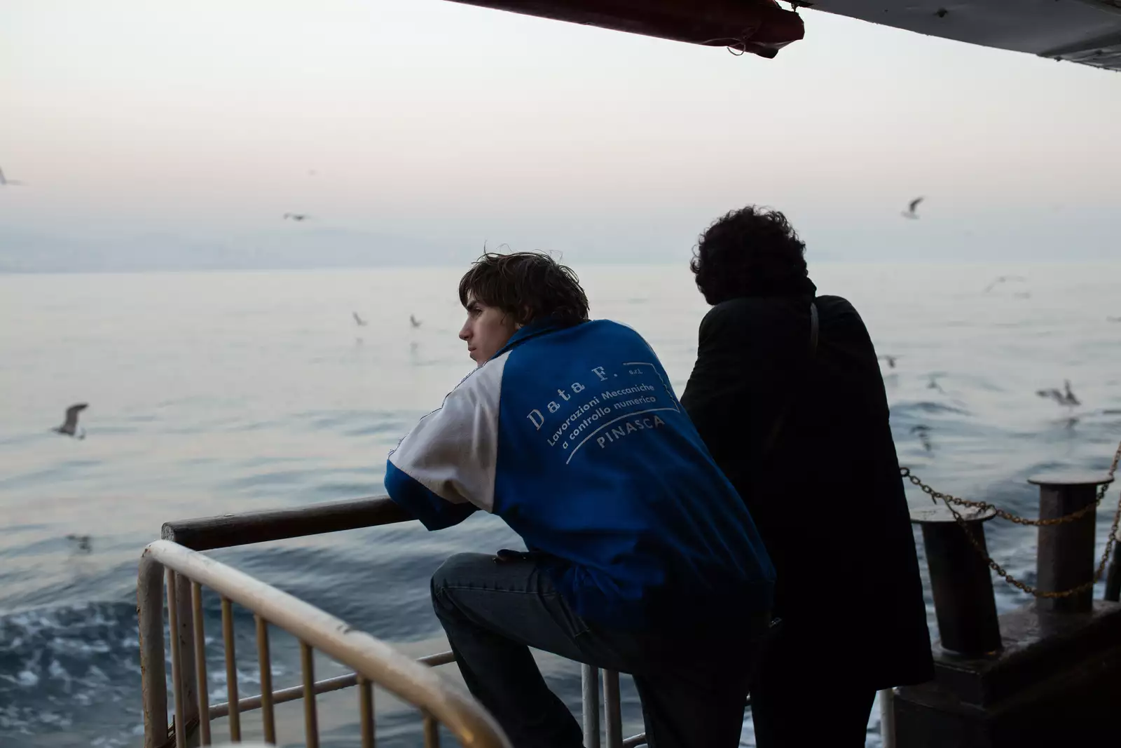Still from Crossing. Two characters with their backs turned, stood by railings looking out at sea.