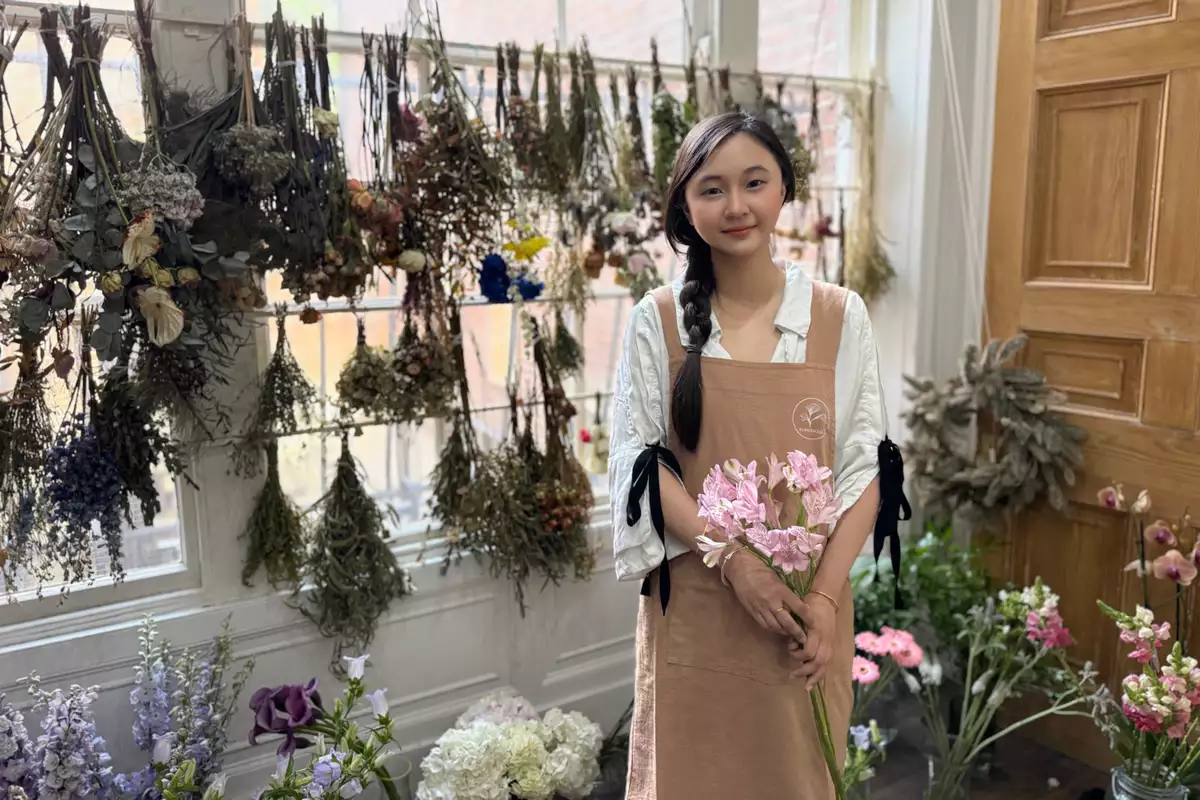 Kiki smiling in her art studio, wearing a light brown apron, holding flowers. There are many bunches of flowers behind her.