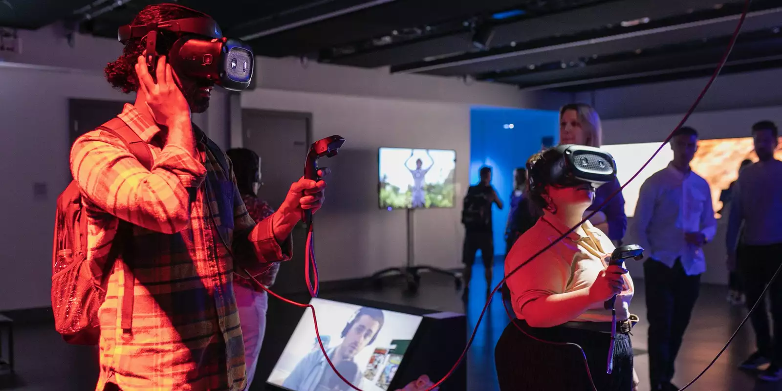 A man and a woman standing in a darkened art gallery, each wearing a Virtual Reality headset and holding a black controller. They are playing a game by the artist Rachel Maclean.