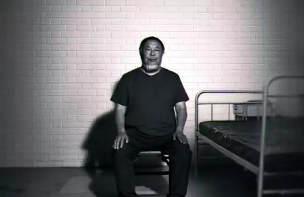 Black-and-white image of Ai Weiwei sitting on a chair beside a simple metal bed against a brick wall.
