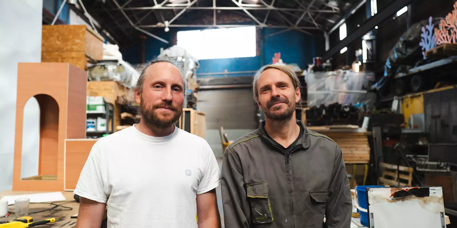 Two members of Studio Tonto stood in a Warehouse filled with equipment. One is wearing a white t-shirt and the other is wearing a grey jacket.