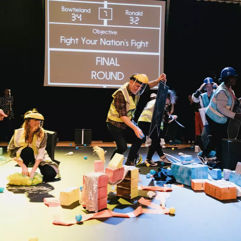 People dressed in safety gear and helmets are actively participating in a theatrical game involving colourful props and foam blocks. A large screen in the background displays the objective "Fight Your Nation's Fight" for the final round, with team scores for Bowtieland and Ronald.