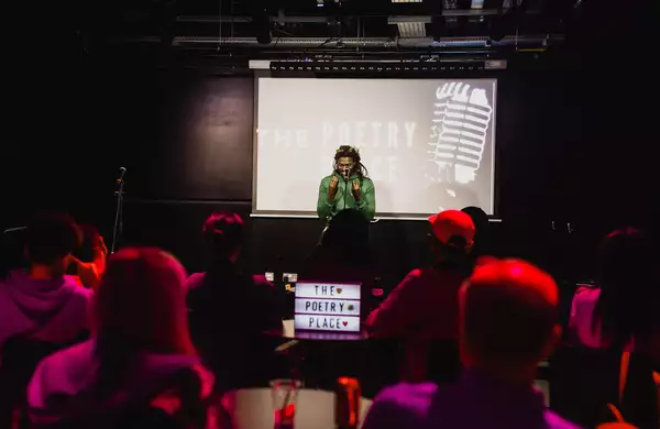A poet performs on stage at a spoken word event as an audience watches in a dimly lit venue. There is a screen behind them which says 'poetry place'.