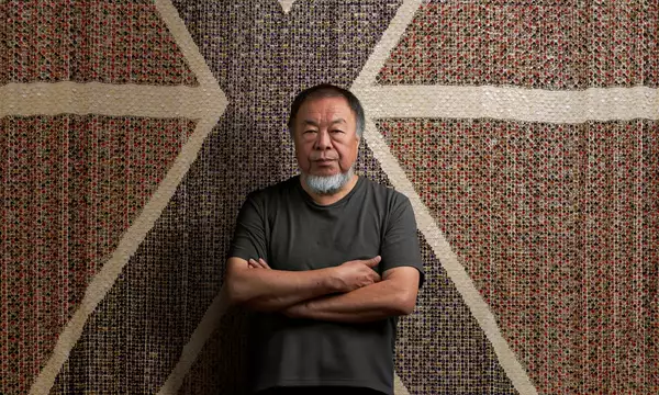 Ai Weiwei stood with arms folded wearing a black t-shirt, in front of a giant flag made of buttons