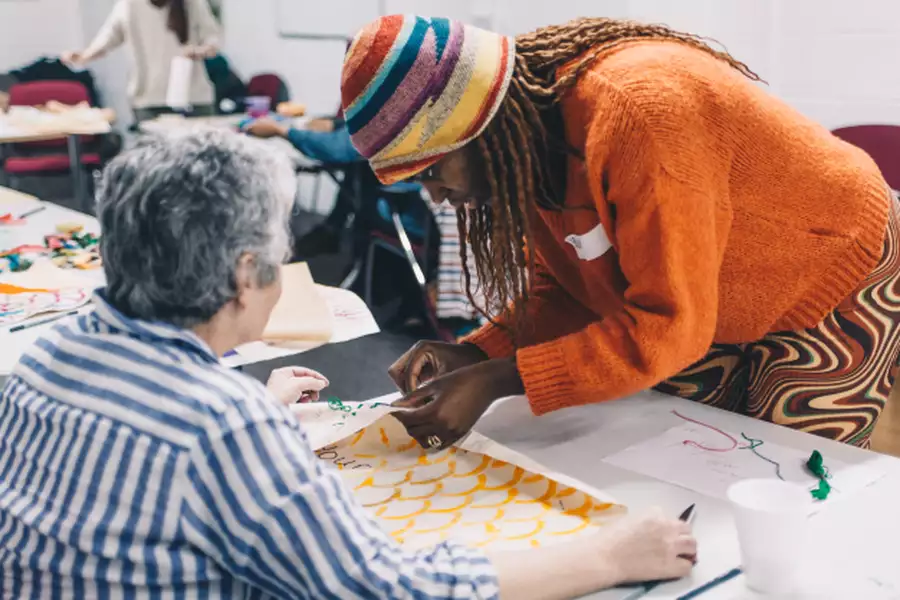 a tutor with a colorful stripy had and blonde braids helps a lady with grey hair with her textile work during a quilt making workshop