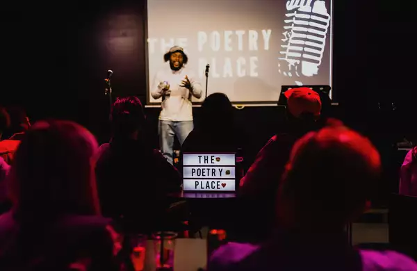 A performer is on stage in the Social with the words 'The Poetry Place' projected on the screen behind them. There is a lit-up sign that says 'The Poetry Place' in front of the stage. The room is dimly lit.