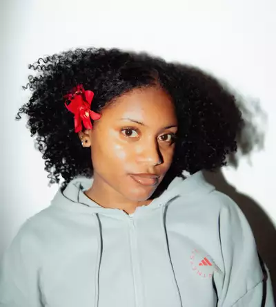 Ella Mayamothi Sommeil wearing a light grey Adidas zipped hoodie and a red flower in her hair, on a white background