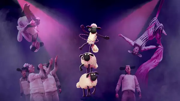 Performers in sheep costumes execute acrobatic stunts and aerial silk acts under dramatic pink lighting. In the center, three animated sheep from "Shaun the Sheep" balance in a tower, with the top one hula-hooping.