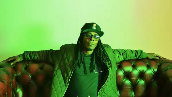 Diligent Fingers sat on a sofa with their arms stretched out. They are wearing a black hat and the lighting is yellow.