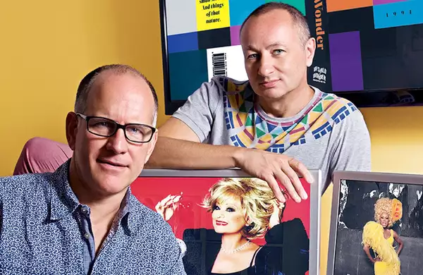 Two people posing with framed photos of performers against a yellow wall