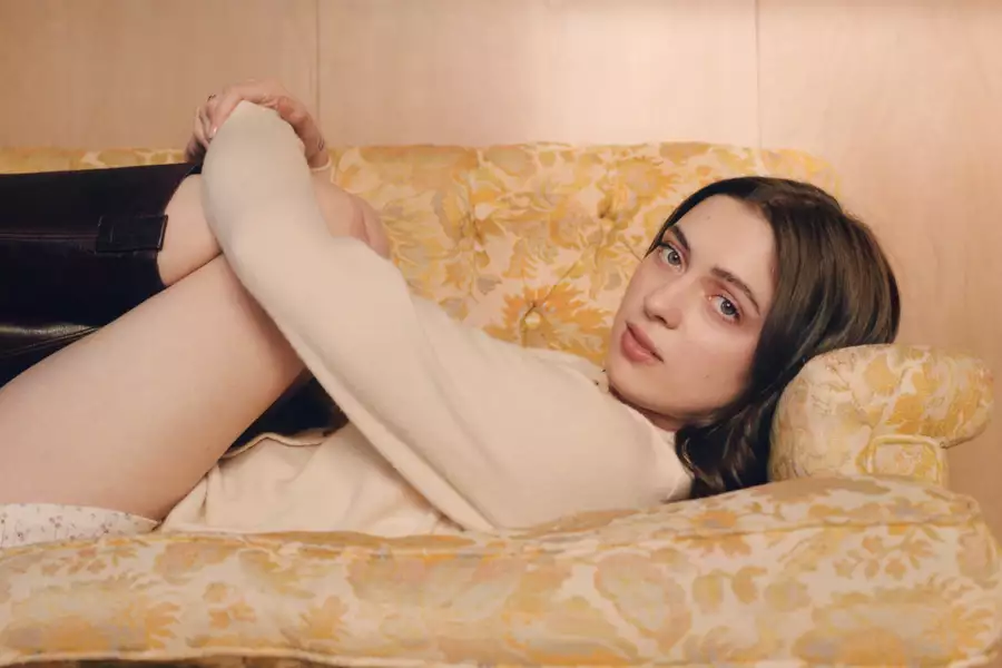 Lizzy McAlpine lying on a yellow and white patterned couch, hugging her knees and looking at the camera