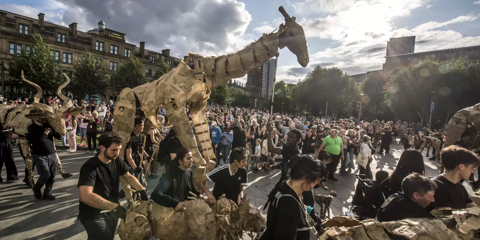 Performers parade life-sized cardboard animal puppets through a city square, watched by a large crowd on a sunny day.