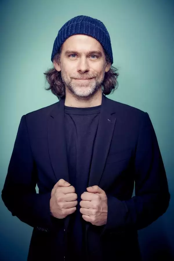 Headshot of Bryce Dessner wearing a black suit jacket, blue beanie and smiling
