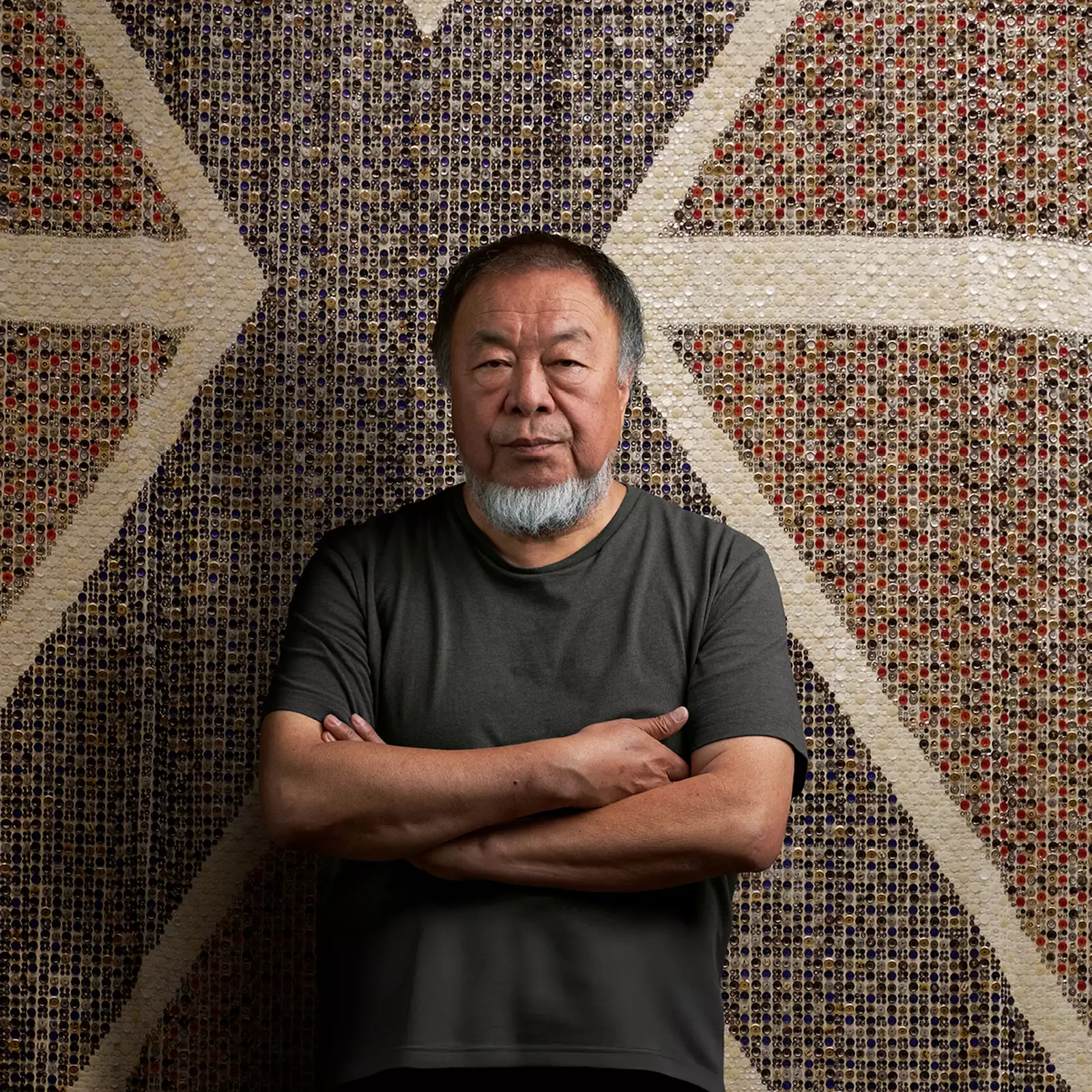 Ai Weiwei stood with arms folded wearing a black t-shirt, in front of a giant flag made of buttons