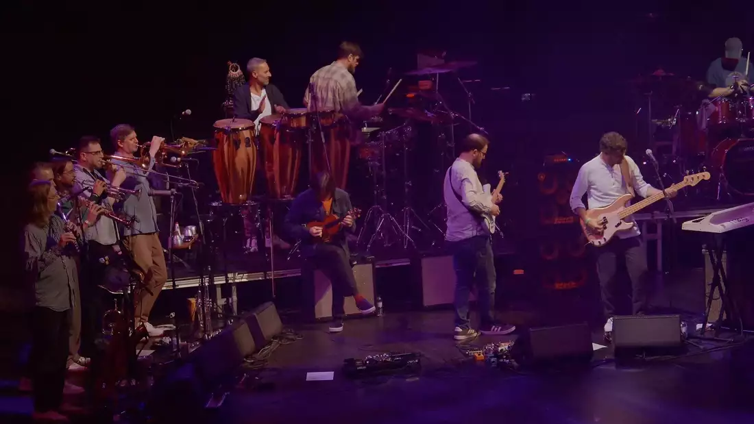 The bad Snarky Puppy performing on stage, with brass players to the left of stage, drummers at the back and guitarists centre and right.