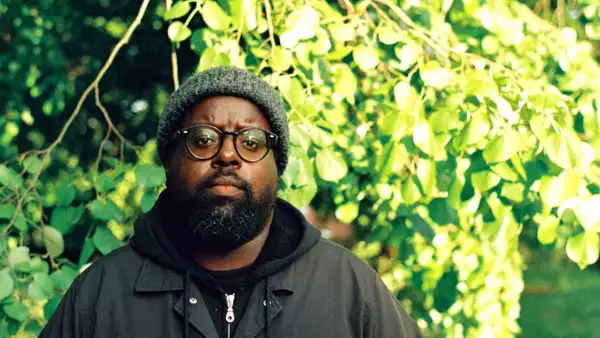 Daudi wearing glasses and a grey beanie stood next to a bush