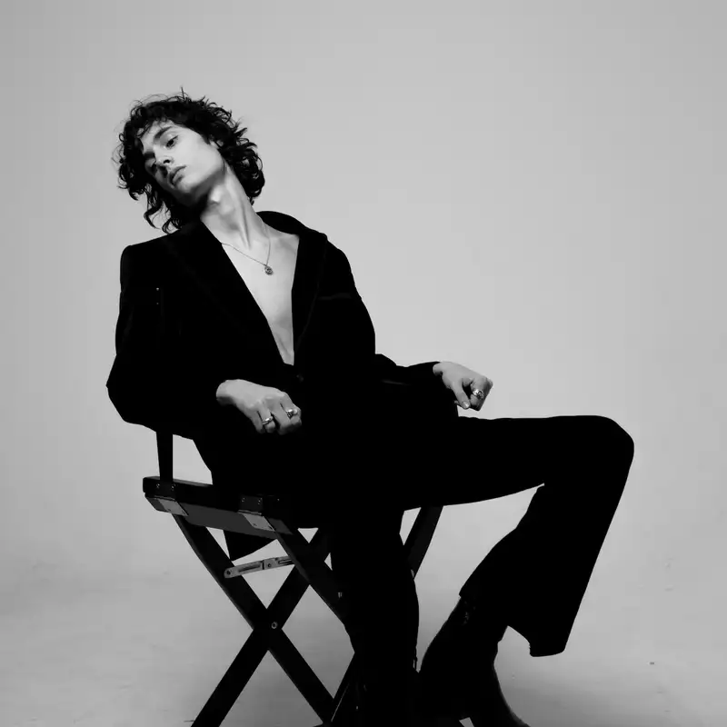 Black and white photo of sombr wearing a suit sitting on a chair