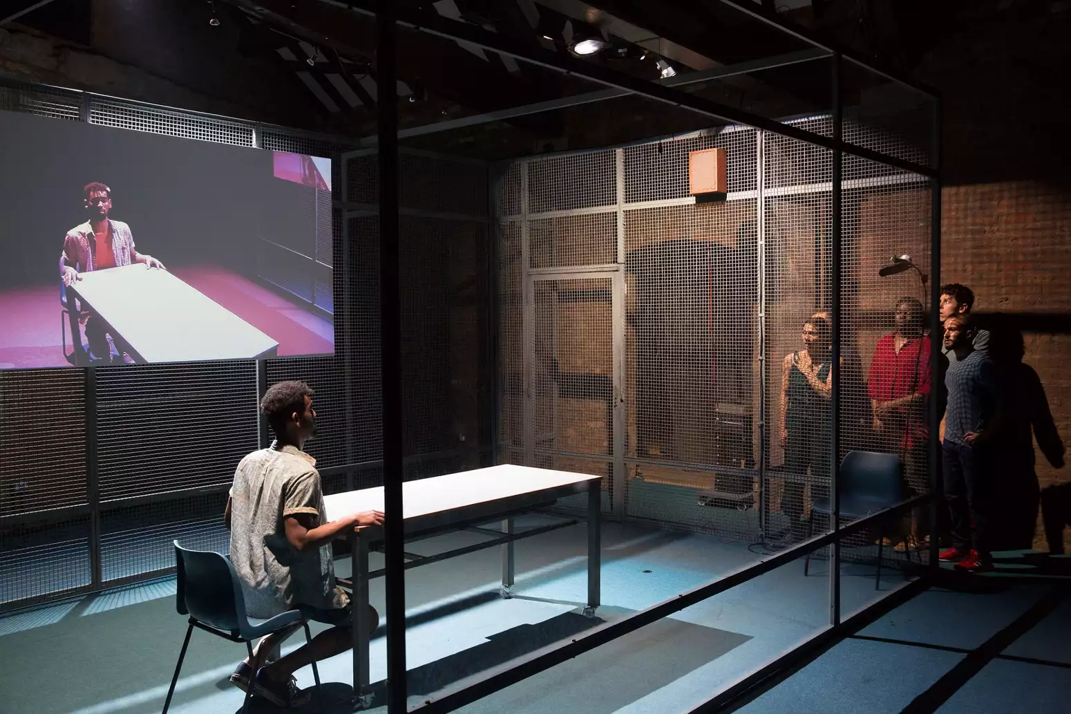 A man sits at a table in a caged room while a group of people look on from outside. An image of the man at the table is projected onto a screen behind him.