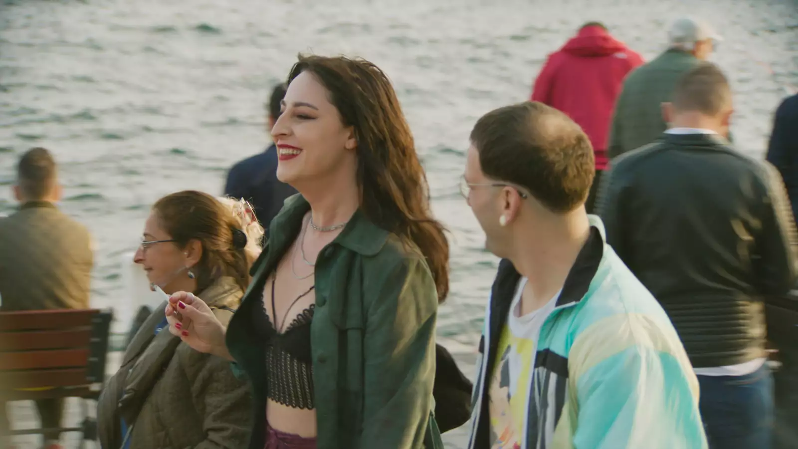 Still from Crossing. Two characters chatting and smiling in a crowd of people stood by the sea. One is smoking.