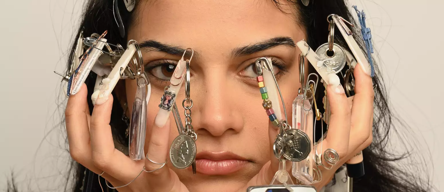 A person with decorated nails holding their face, featuring various keychains and trinkets attached to their nails.