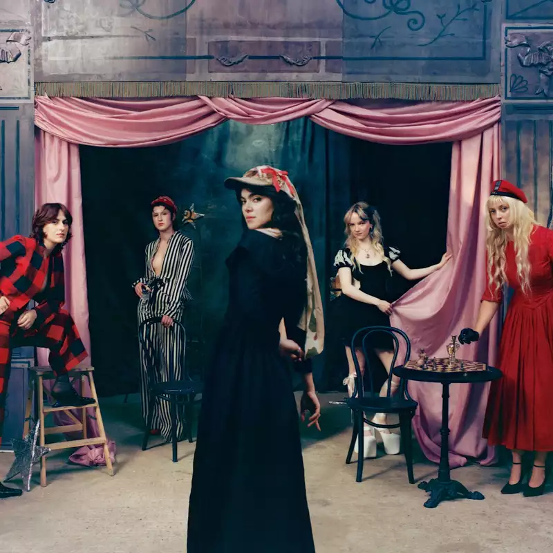 Five people from The Last Dinner Party in theatrical costumes pose on a staged set with pink curtains, including figures in black, red, and striped outfits around a small table with a chessboard.