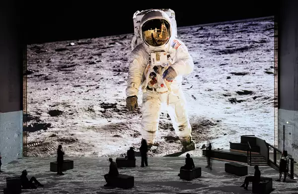 People sit and stand in a dark immersive exhibition space with a massive floor-to-ceiling projection of an astronaut walking on the moon. The lunar surface image extends across the floor and walls, creating a sense of being on the moon.