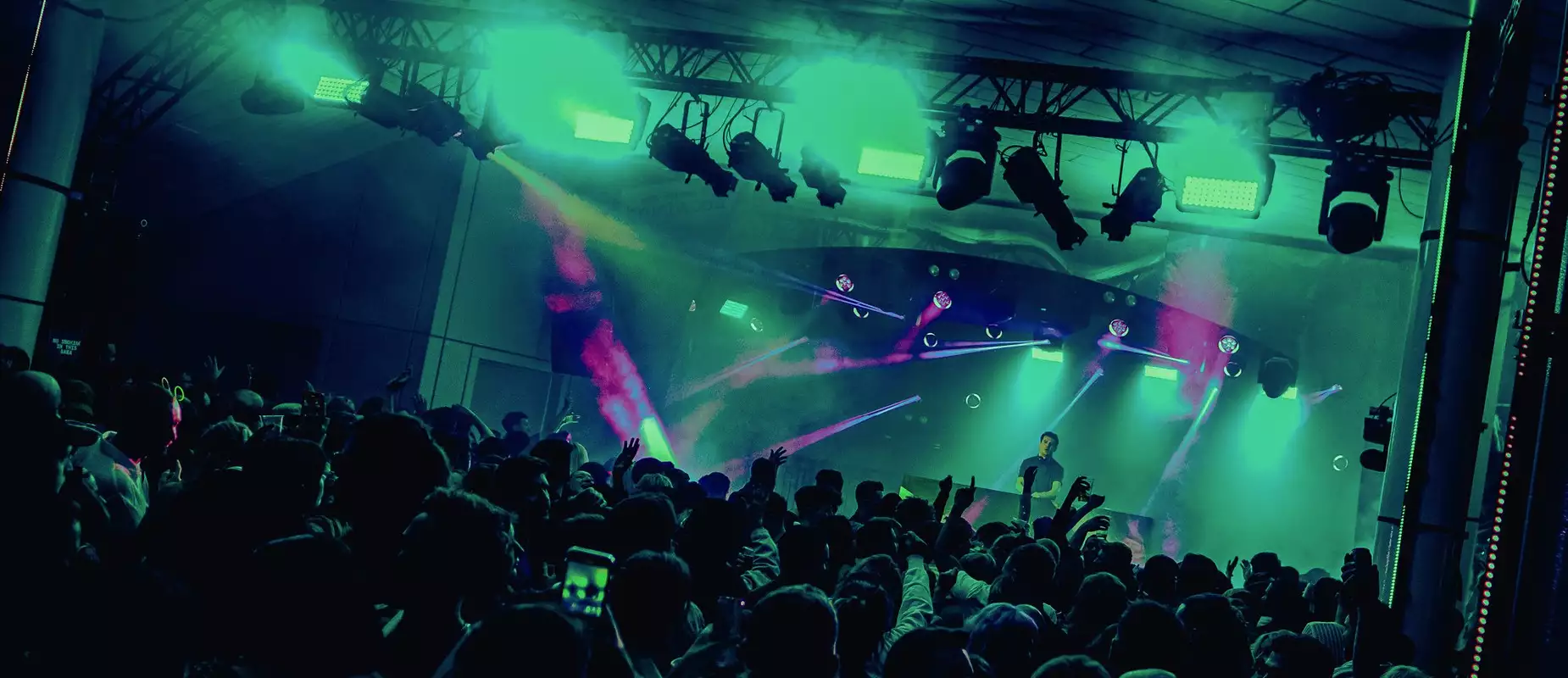 A DJ set on Festival Square from MIF23, showing a large crowd of people dancing and filming a DJ on stage. The lighting is hazy green and there are purple strobe lights.