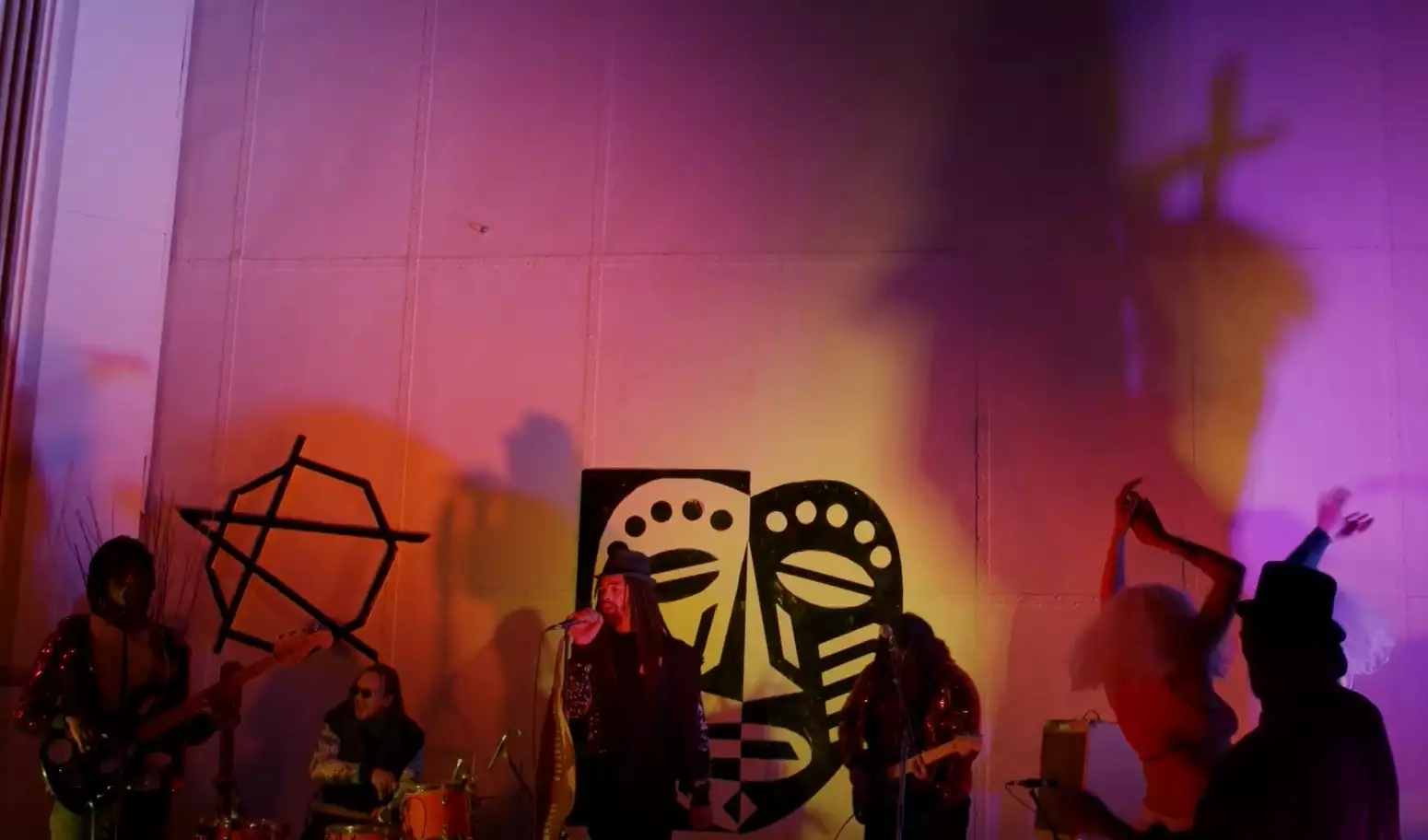 A band playing in front of a white wall. There is low level lighting so the figures are hard to make out, there are shadows on the wall. From left to right, bassist and vocalist, drummer, vocalist, guitarist, then two dancers - one wearing a top hat.