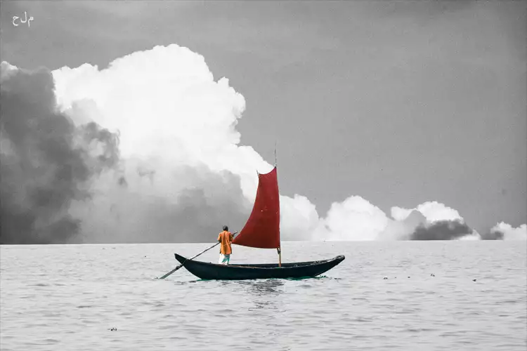 A calm sea with puffy grey and white clouds on the horizon, in black-and-white. Floating on the water’s surface, in colour, is a wooden boat with a single red sail. An old man stands in the boat wearing an orange shirt and holding a large oar. In the top left-hand corner are the letters: م ل ح