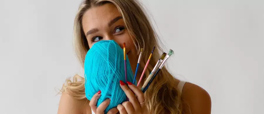 A woman posing with a bright blue ball of wool and paintbrushes