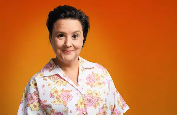 Susan Calman smiling and wearing a flowery shirt on a bright orange background