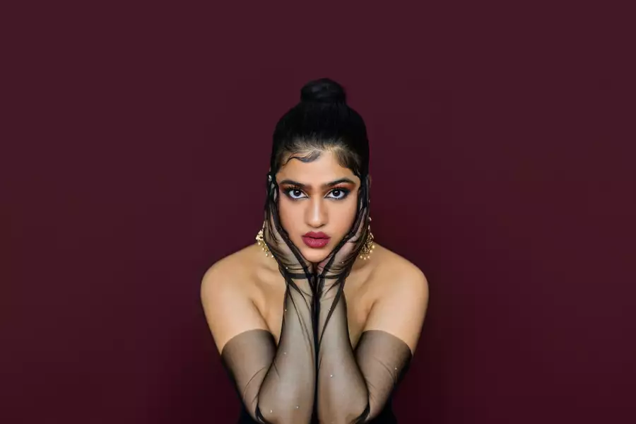A woman poses confidently against a deep maroon background, gazing directly into the camera with intensity. Her hair is styled in a sleek top bun, and she wears dramatic makeup with bold eyeliner and deep pink lipstick. She rests her face in her hands, which are adorned with sheer black gloves decorated with subtle sparkles. Gold earrings peek out from behind her fingers, adding a touch of glamour.