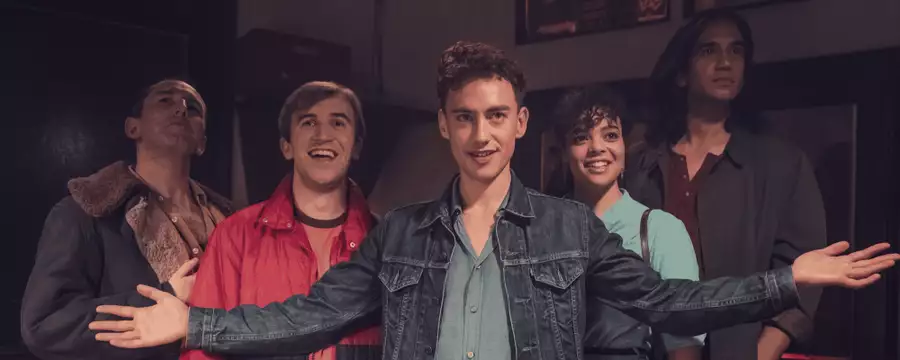 Cast image from It's a Sin showing five friends standing together indoors, smiling and looking upward, with the person in front wearing a denim jacket and holding their arms out wide.