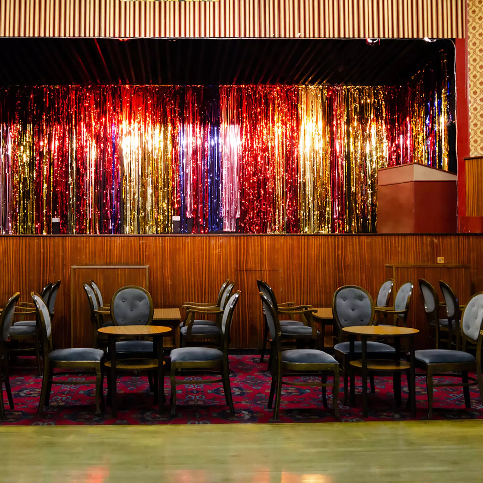 A pub stage with a glittery curtain, blue chairs, tables and a patterned carpeted floor.