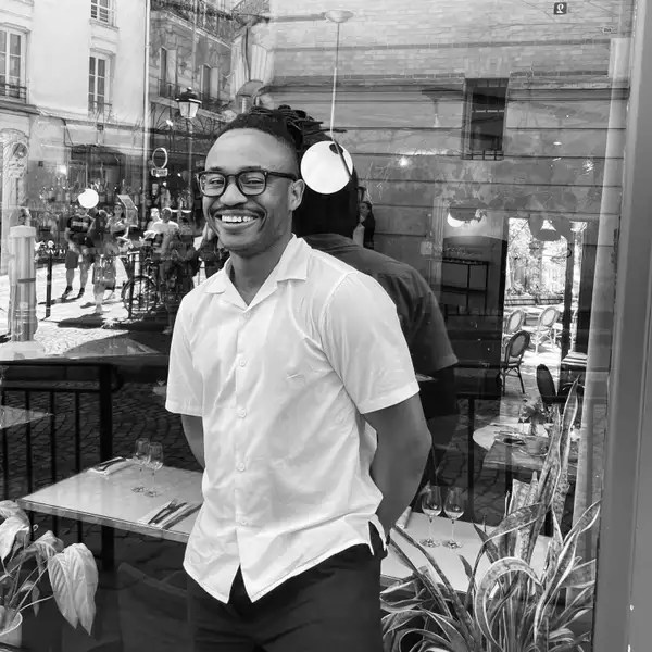 Black and white square photograph of Otega Ajuchi wearing a short-sleeved shirt and smiling