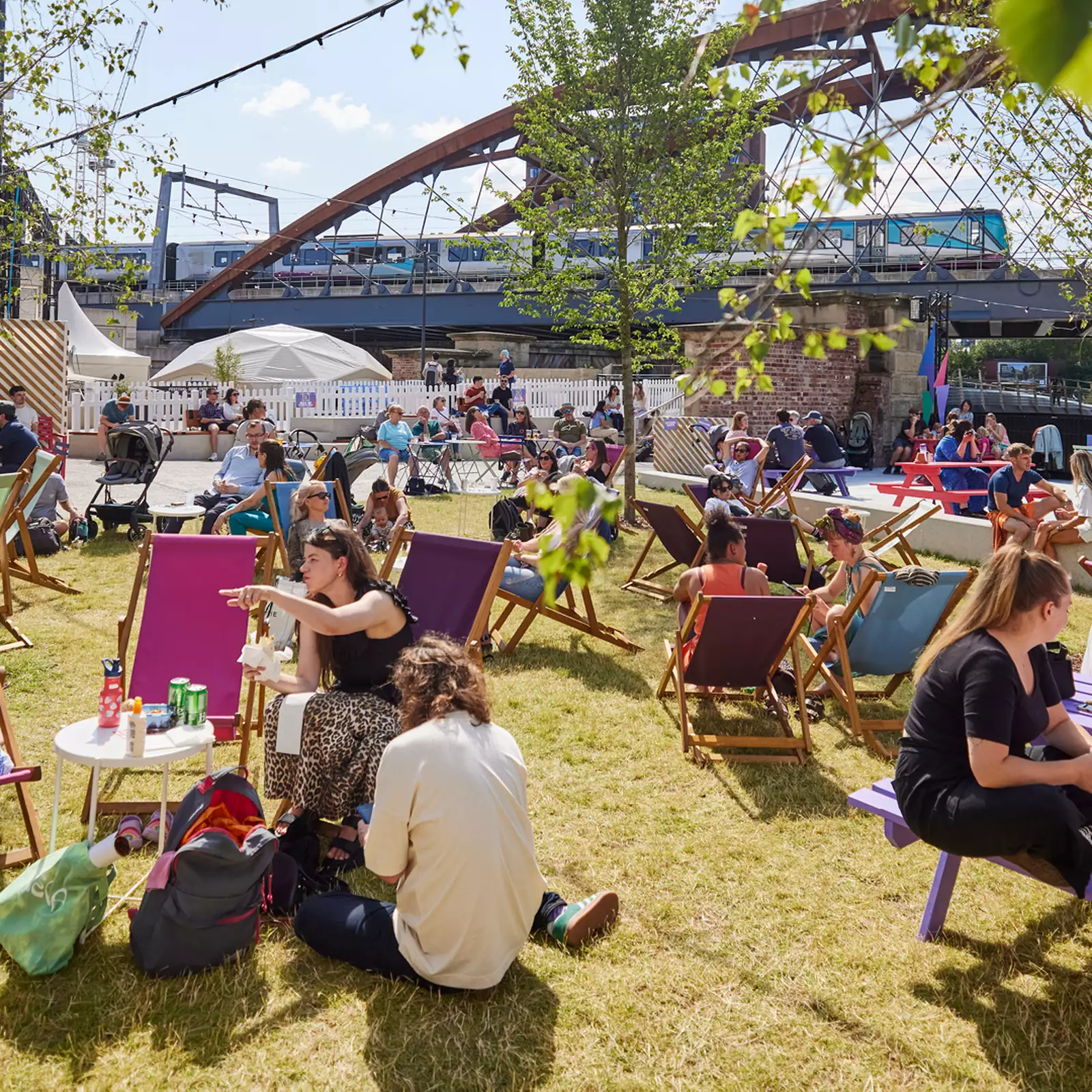Factory Square filled with people sat on the grass and deckchairs, drinking and chatting. The sun is shining.