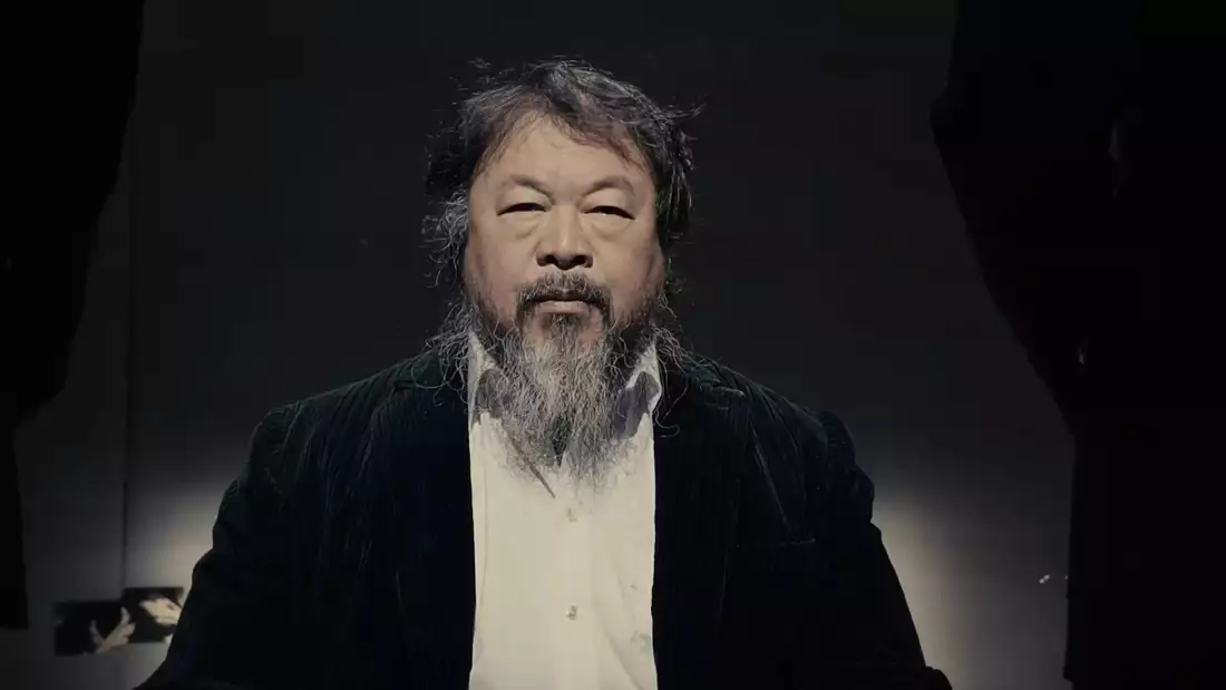 Ai Weiwei wearing a white shirt and black jacket in a dark room