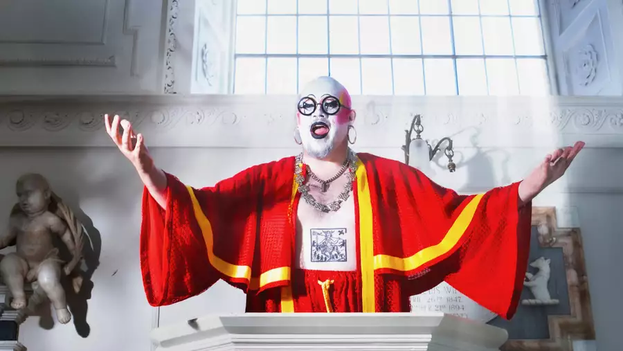 Fatt Butcher in makeup and a bright red robe stands at a pulpit with arms outstretched in a sunlit church.