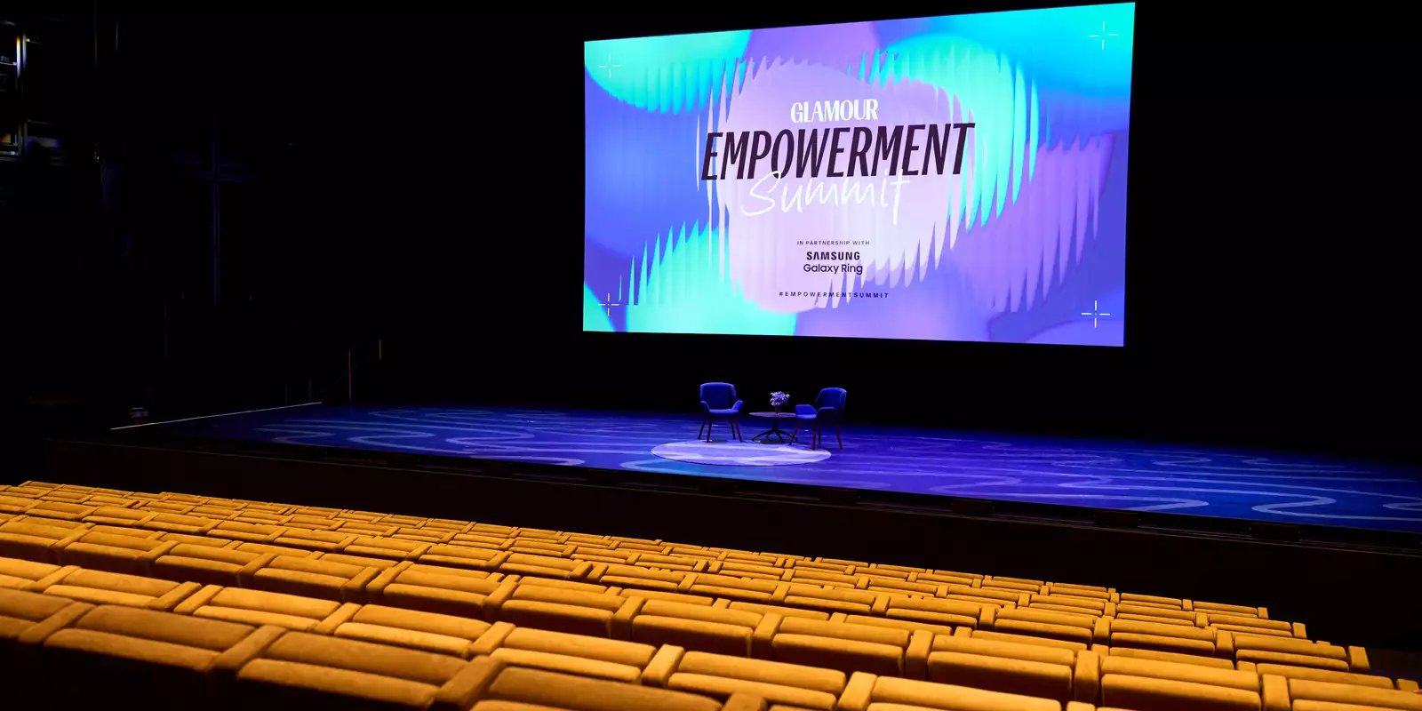 Empty seats in the Hall. The stage has two empty chairs on it ready for an event. The screen behind says 'Glamour Empowerment Summit'.